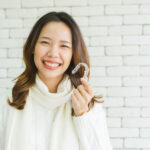 Asian woman smiles while wearing a cream turtleneck sweater and holding her Invisalign aligners