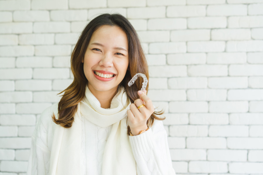 Asian woman smiles while wearing a cream turtleneck sweater and holding her Invisalign aligners